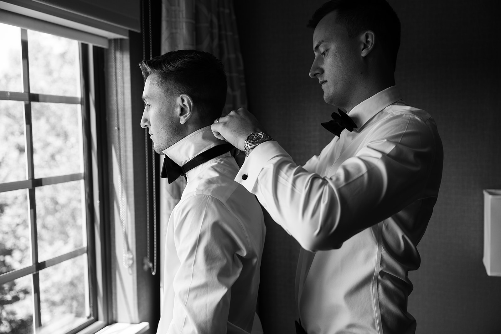 Black and white photo of a groomsman adjusting the groom's collar as he prepares by a window.